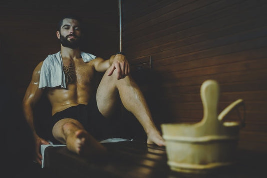 Un uomo seduto su una panca in una sauna con accanto un secchio di legno.