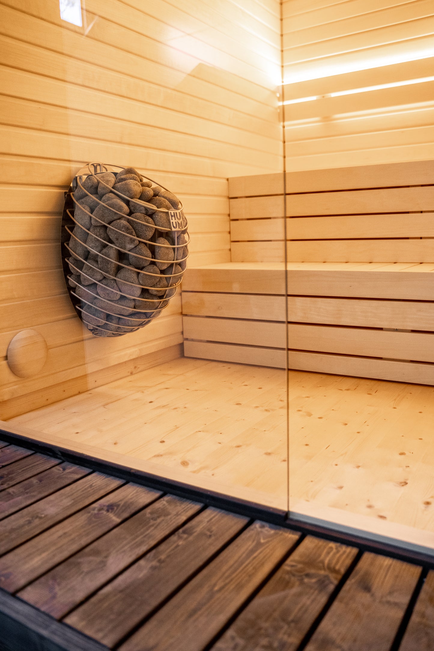 Sauna LUMI con cesto in pietra e parete in vetro.